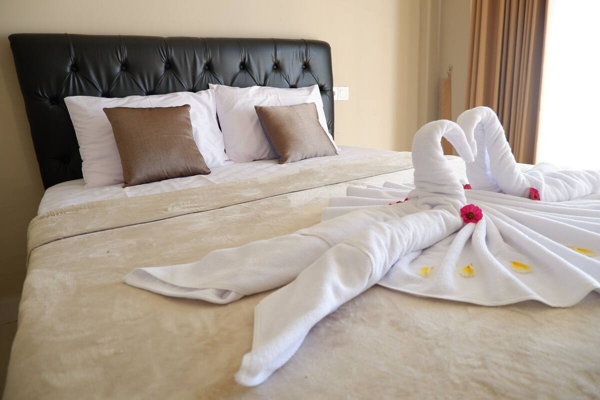A neatly made bed with a black headboard, white and brown pillows, and a white towel arranged in a swan shape with a pink flower.