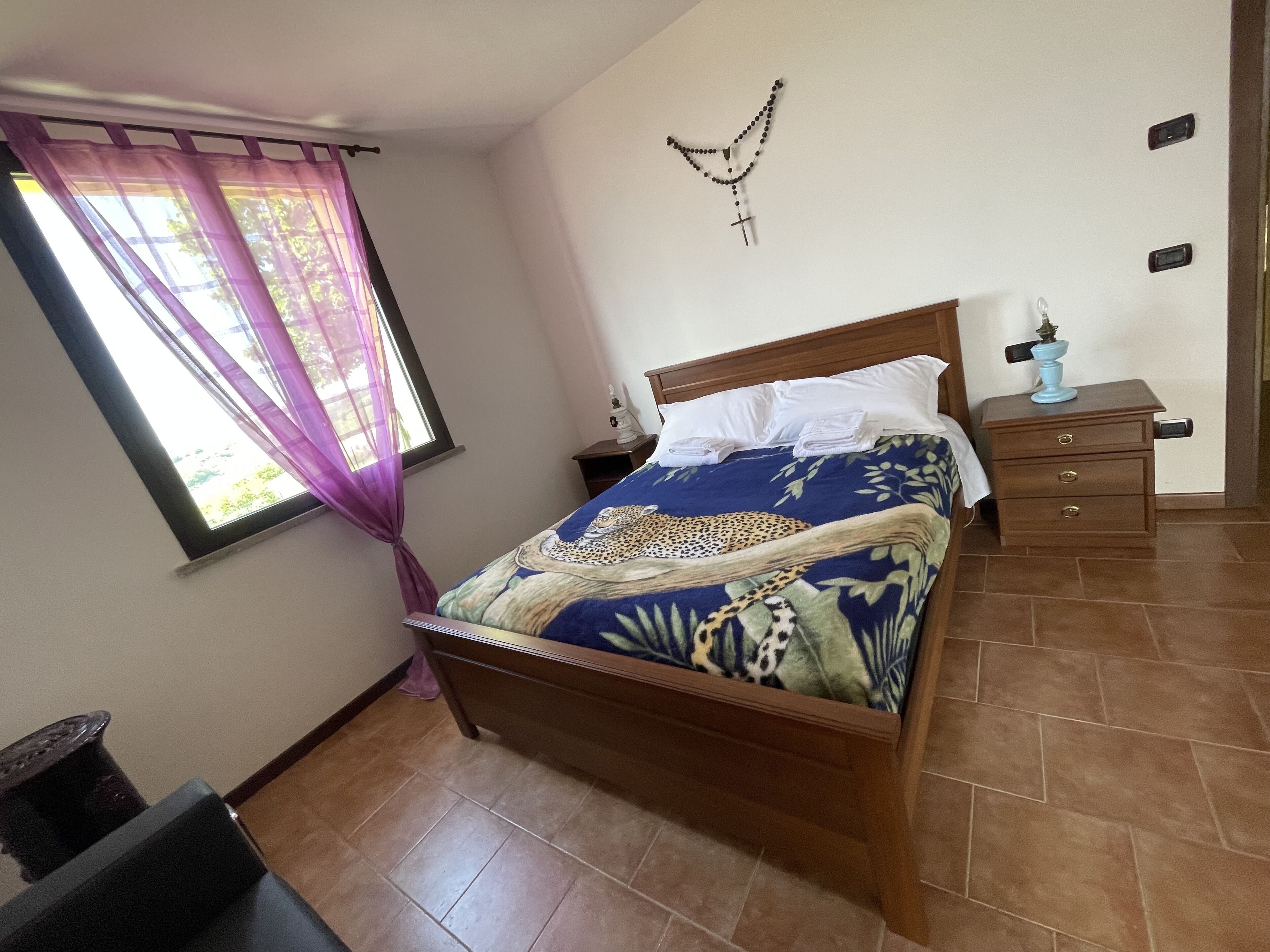Un dormitorio con cama, mesita de noche, un crucifijo en la pared y ventana con cortinas.