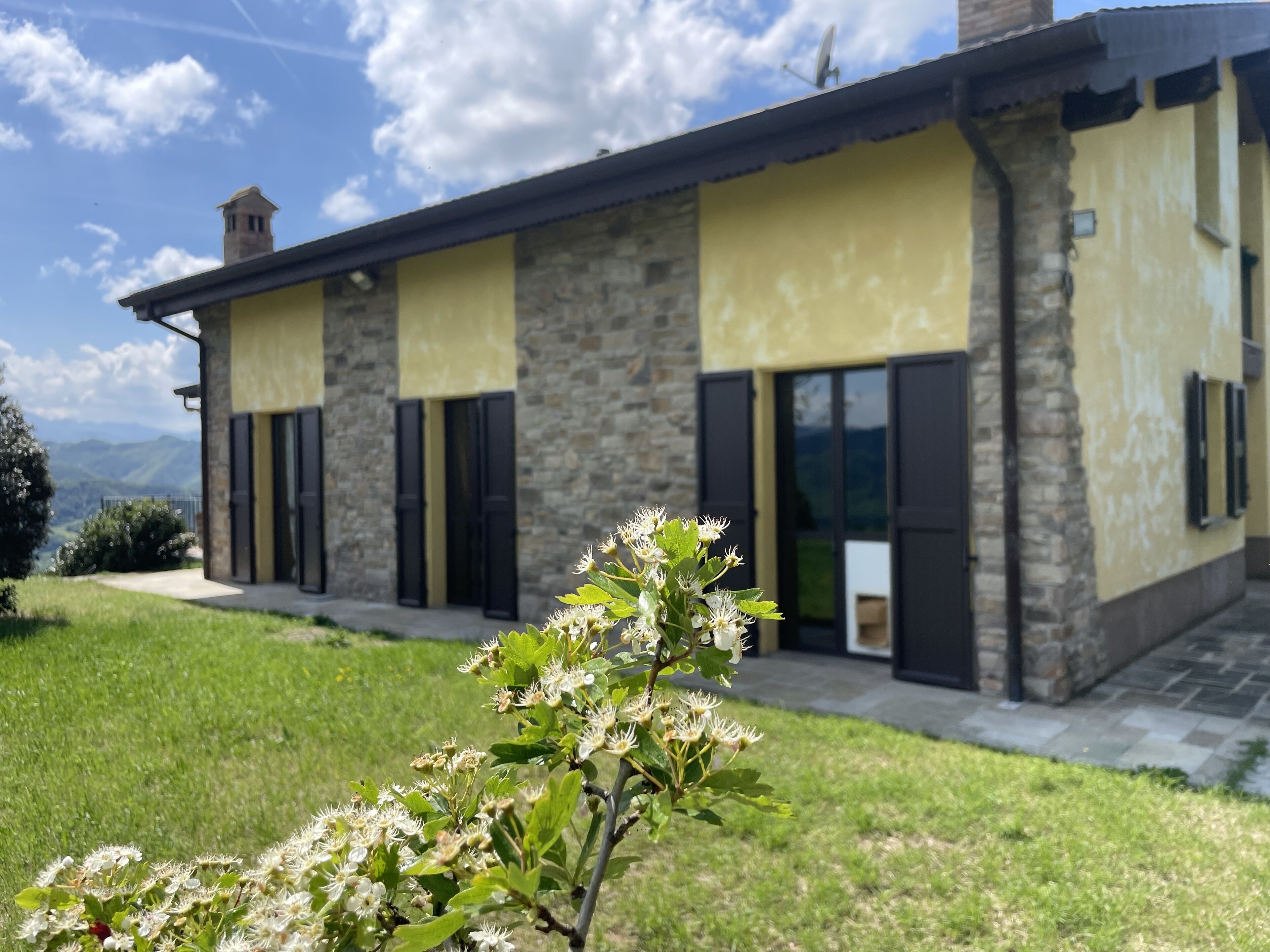Ein Steinhaus mit gelber Fassade, schwarzen Fensterläden und Blick auf die Berge.