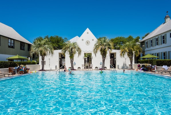 A swimming pool with a clear blue water surface, surrounded by palm trees and a building with a clock on the facade.