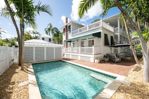 Une piscine entourée d’une clôture blanche, avec des palmiers et une maison à deux étages dotée d’un balcon.
