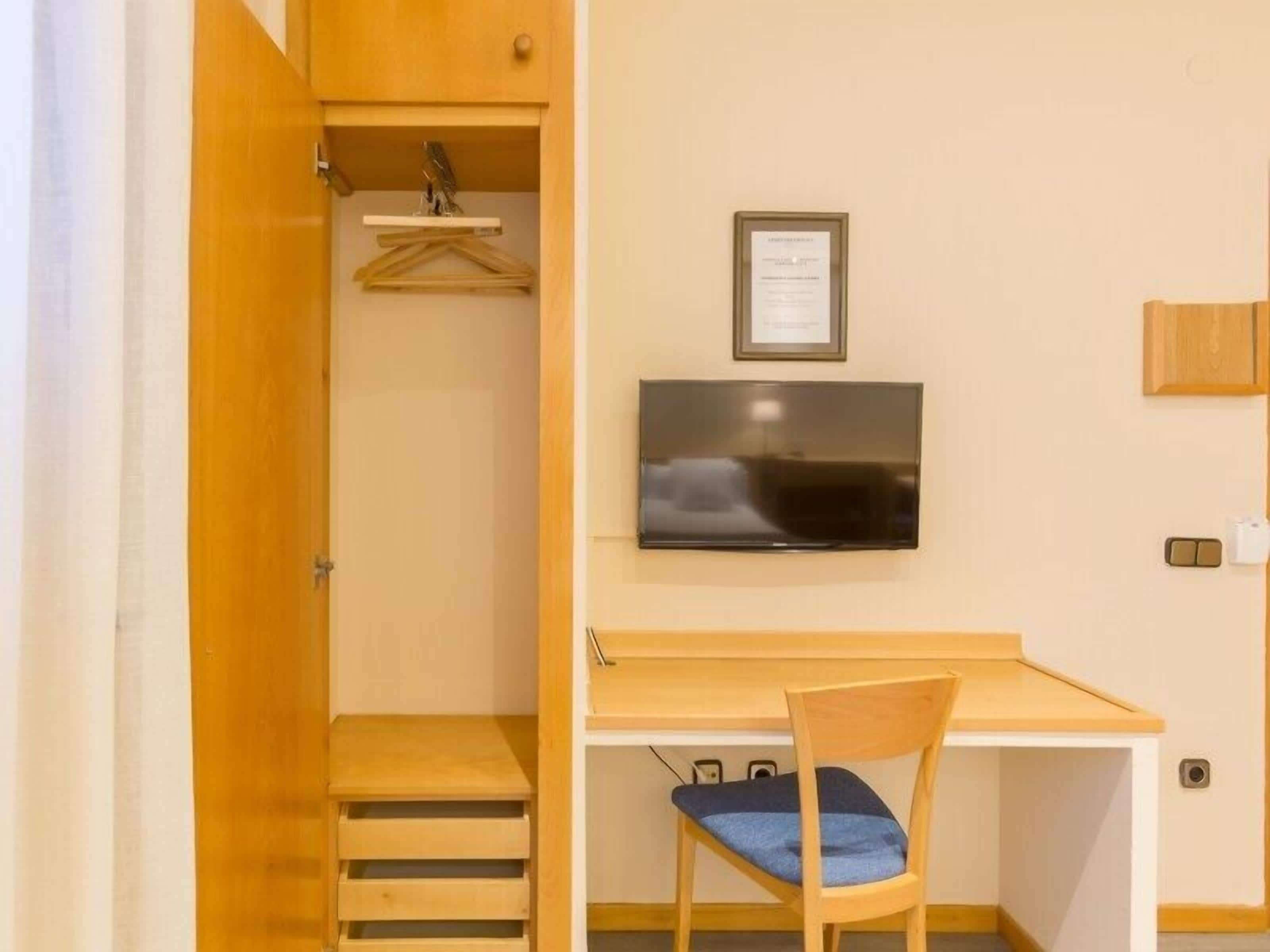 Una habitación compacta con un escritorio de madera, una silla, un televisor montado en la pared, un estante pequeño con un objeto colgando y una puerta que da a otra habitación.
