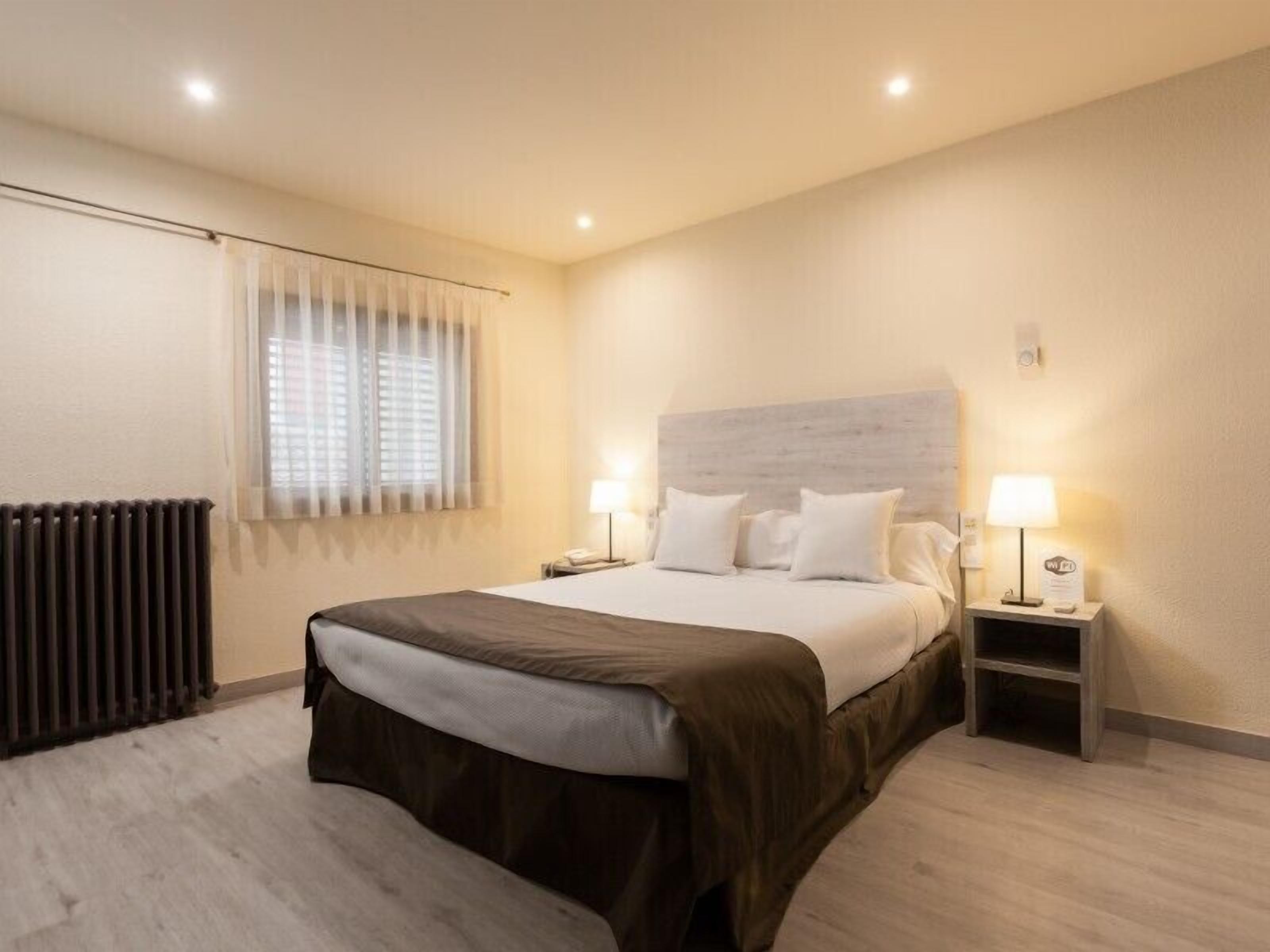 Habitación de hotel con una cama grande, dos mesitas de noche con lámparas, un radiador y una ventana con persianas.