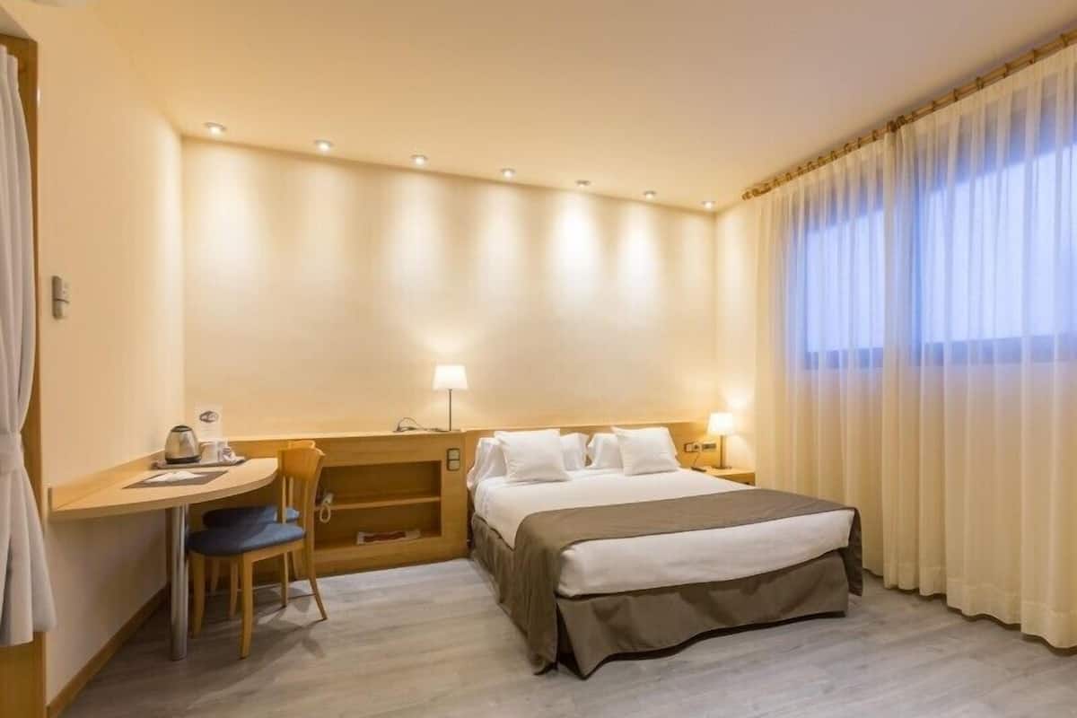 A hotel room with a bed, a desk, a chair, and a window with curtains.