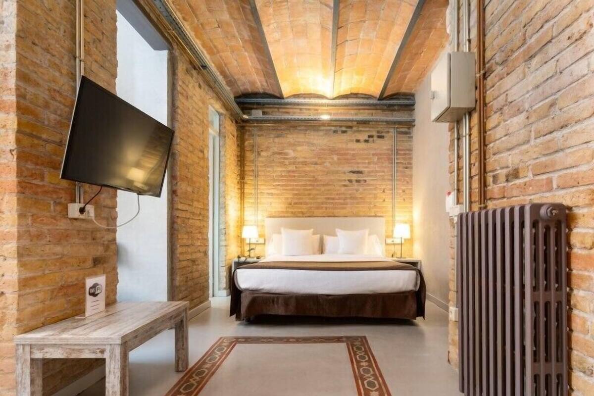 A hotel room with exposed brick walls, a large bed with white linens, a wooden bench, a flat-screen TV mounted on the wall, and a radiator.
