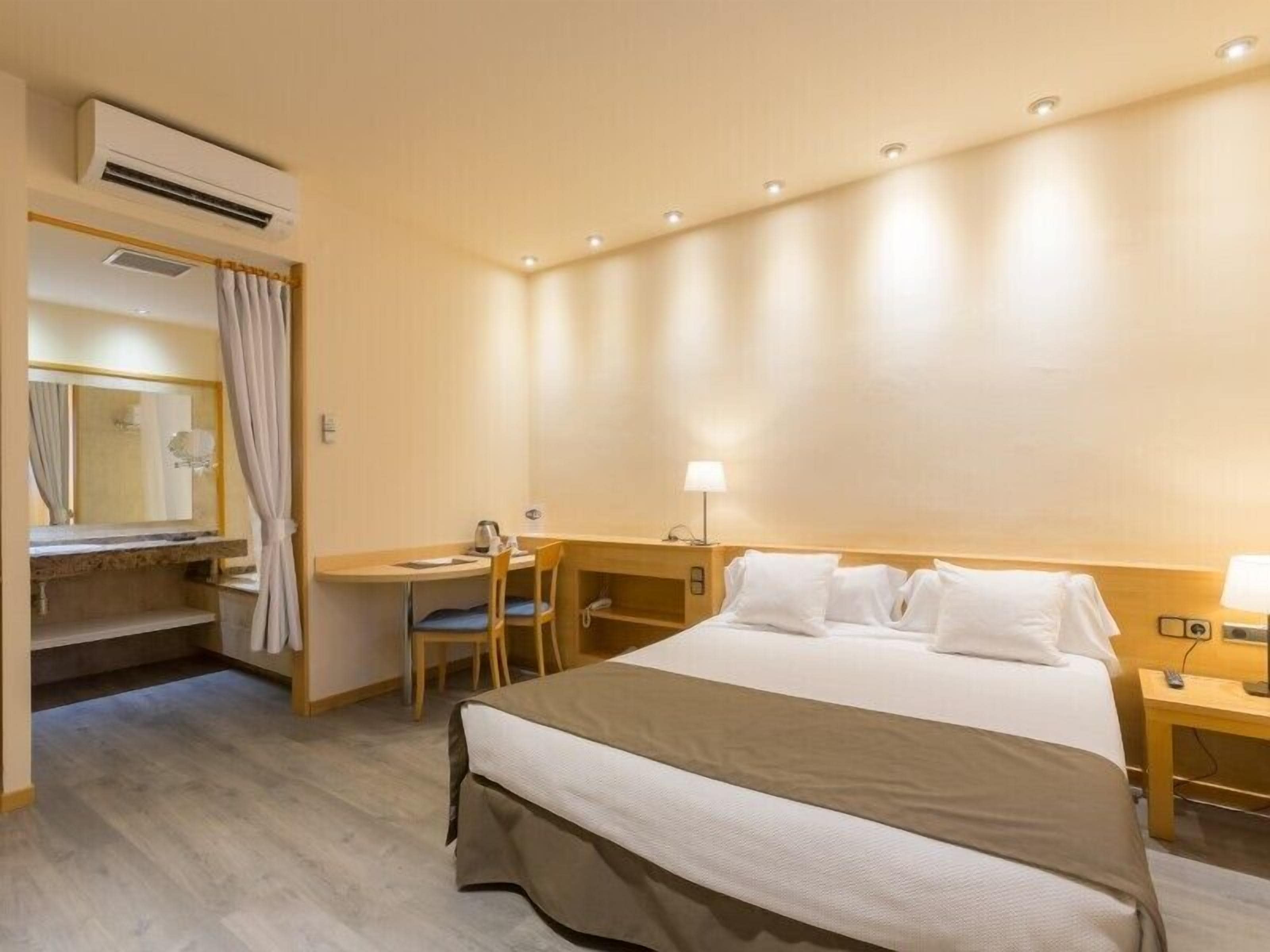 Una habitación de hotel con una cama, un escritorio, una silla, una lámpara y un aire acondicionado.