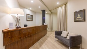 A reception area with a wooden desk, a lamp, a framed picture, and a grey sofa. - Hospedium Apartamentos DV (Barcelona)