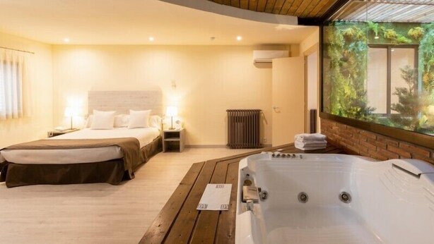 A modern hotel room with a wooden ceiling, a large bed, a separate smaller bed, a hot tub, and a view of greenery through a glass wall. - Hospedium Apartamentos DV (Barcelona)