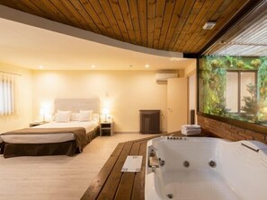 A modern hotel room with a wooden ceiling, a large bed, a separate smaller bed, a hot tub, and a view of greenery through a glass wall. - Hospedium Apartamentos DV (Barcelona)