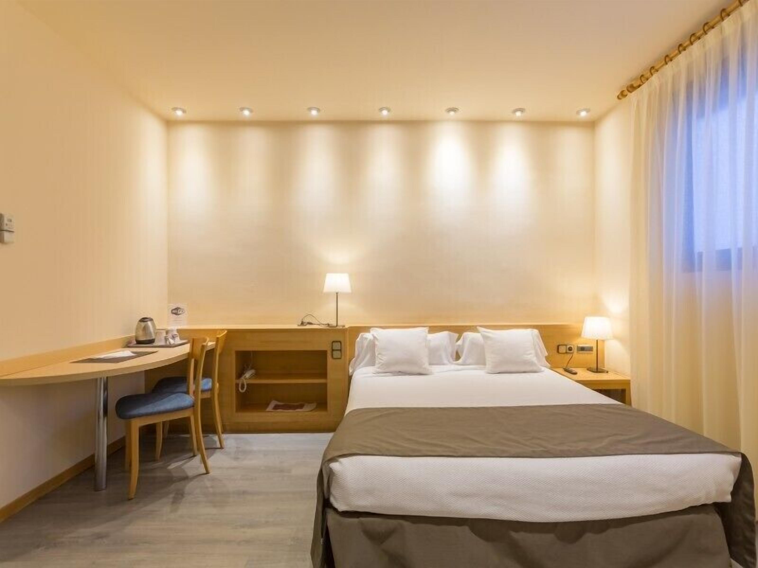 Quarto de hotel com cama, escrivaninha, cadeira, mesa de cabeceira e abajur.