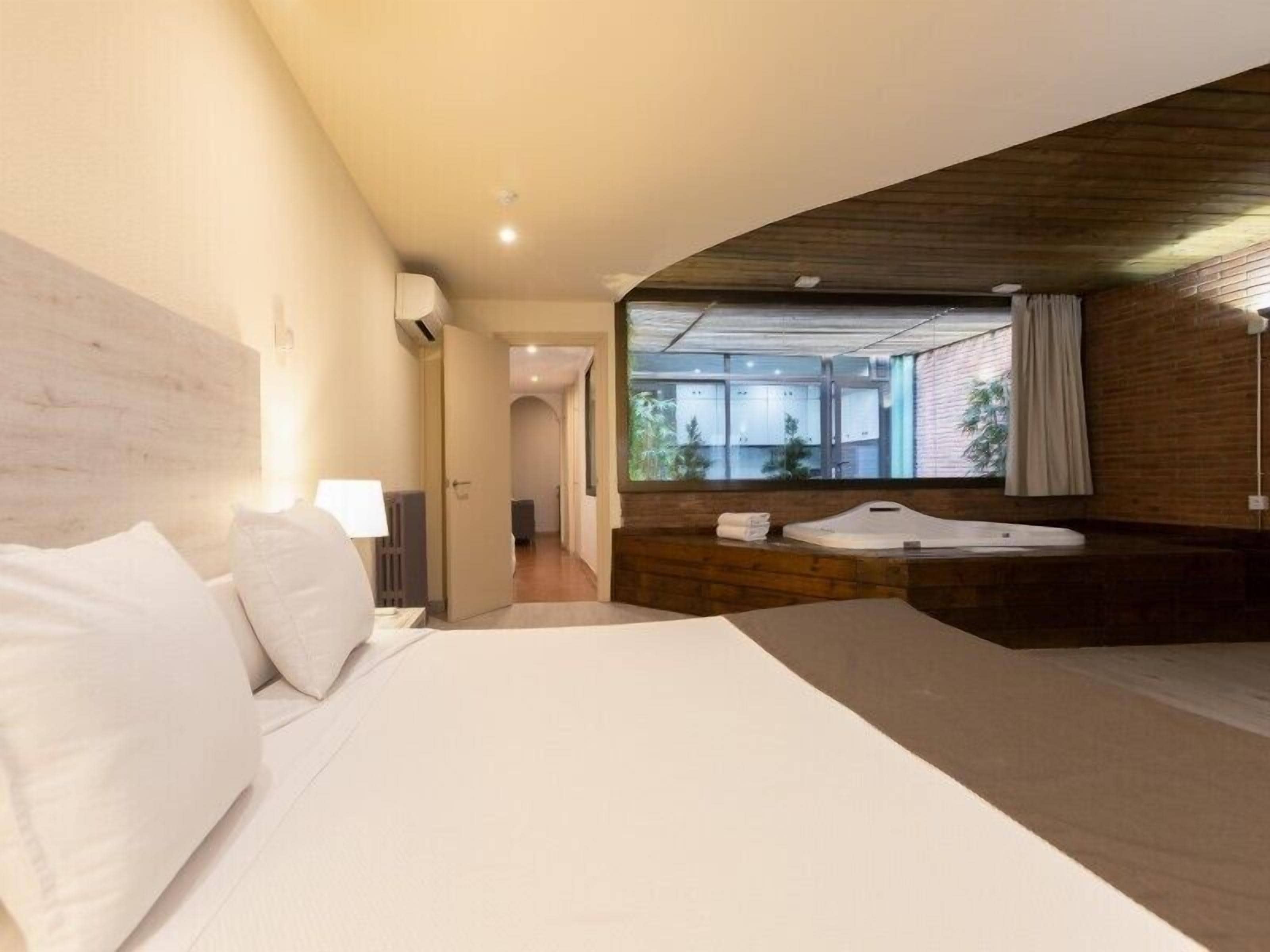 Habitación de hotel moderna con una cama grande, cabecera de madera, bañera integrada y vistas al exterior.