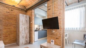 A modern living room with exposed brick walls, a flat-screen TV mounted on a brick pillar, a wooden bench, and a view into a kitchen area. - Hospedium Apartamentos DV (Barcelona)