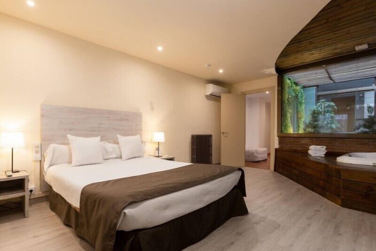 A modern hotel room with a large bed, bedside lamps, a wooden accent wall, and a view of greenery through a window.