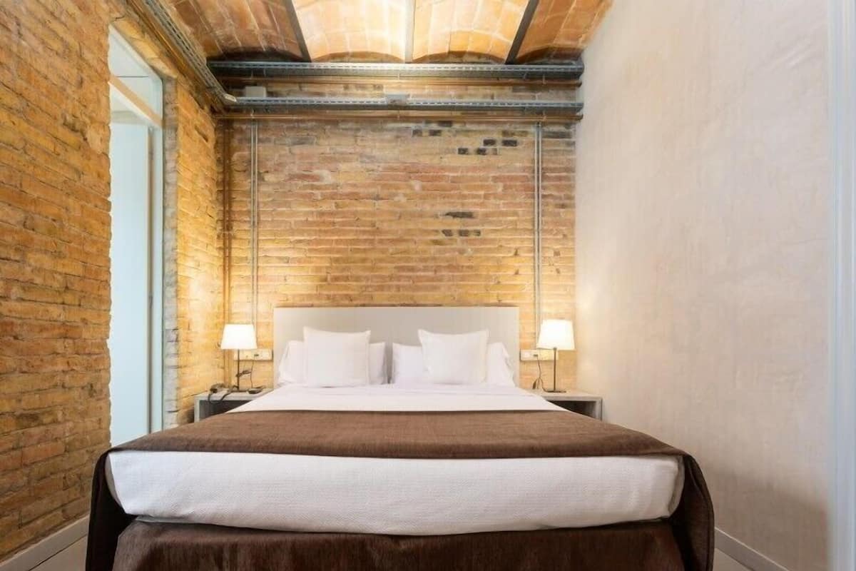 A modern hotel room with exposed brick walls, a large bed with white linens, and two bedside lamps.