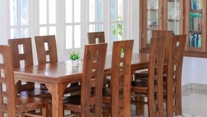 A wooden dining table with six chairs, a small plant on the table, and a bookshelf in the background. - Everest Lodge  (Hulhudhoo)