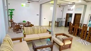 A spacious living area with sofas, armchairs, and a coffee table. There is a dining table with chairs and a potted plant. The room has a high ceiling with recessed lighting and ceiling fans. - Everest Lodge  (Hulhudhoo)