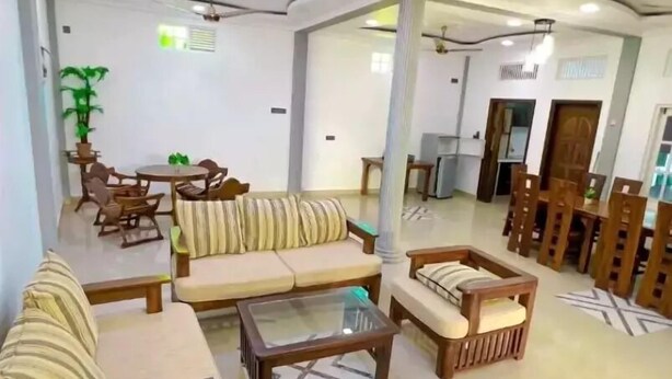 Amplia sala de estar con sofás, sillones y una mesa de centro. Se observa una mesa de comedor con sillas y una planta en maceta. La habitación tiene un techo alto con iluminación empotrada y ventiladores de techo.