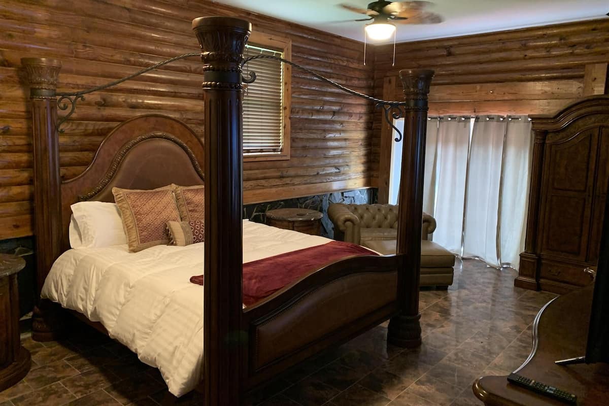 Uma cama com dossel e cabeceira de madeira, uma poltrona estofada, uma mesa pequena e um espelho.