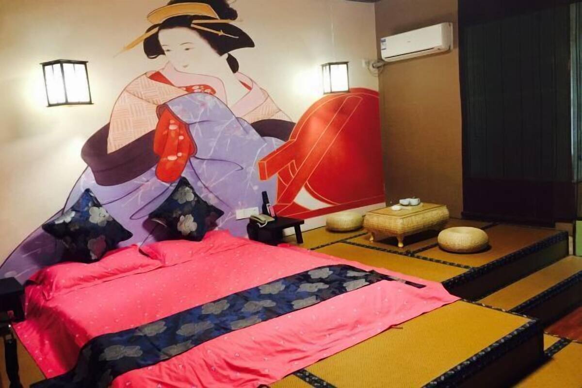 A traditional Japanese-style room with tatami flooring, a low bed with a pink and blue bedspread, a red chair, and a wall painting.