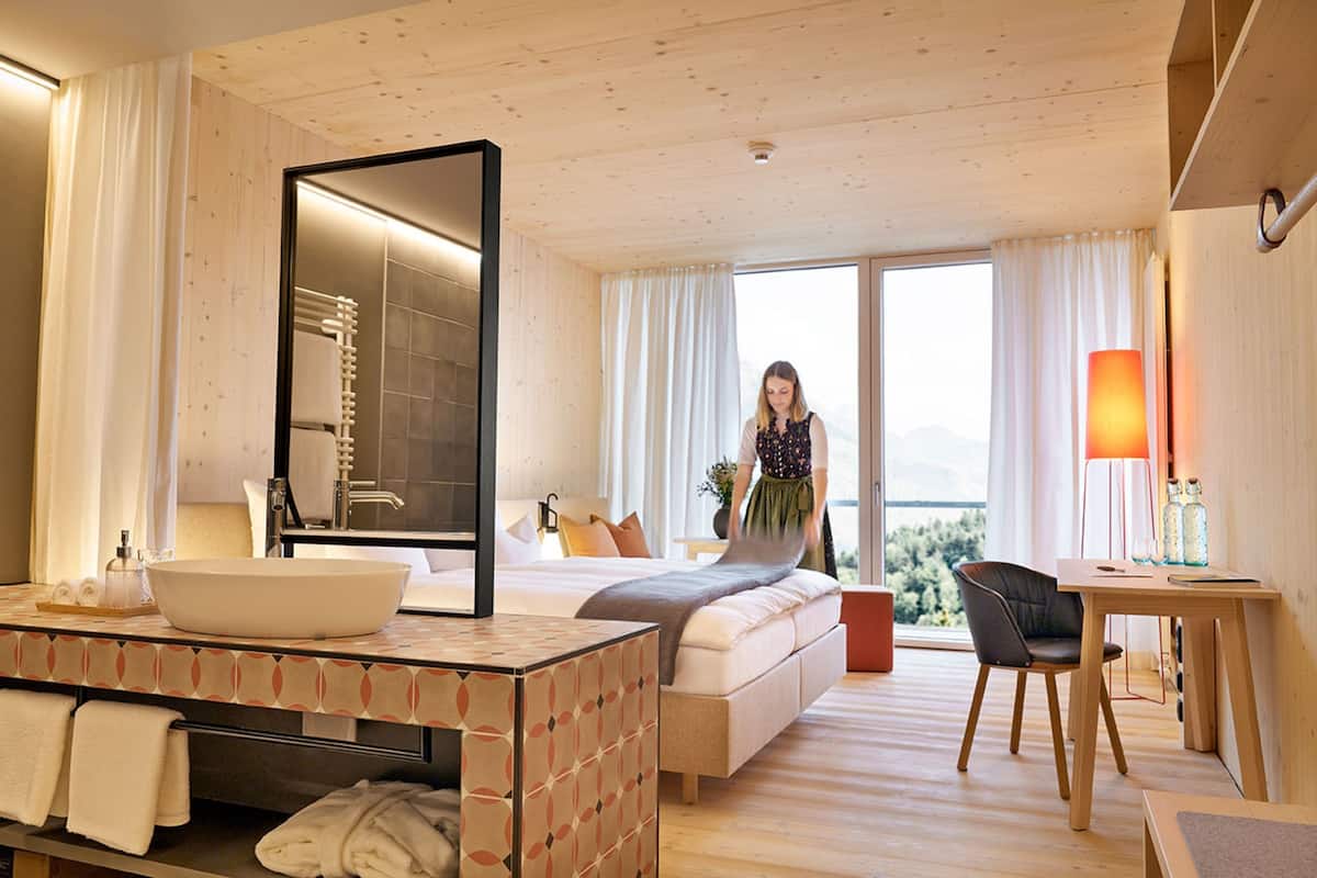 Une chambre d’hôtel moderne dotée d’un grand lit, d’une salle de bain avec un grand miroir, d’un bureau et offrant une vue sur l’extérieur.