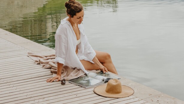 Une femme est assise au bord d’une piscine tranquille, avec une terrasse en bois, un chapeau de paille et un livre.