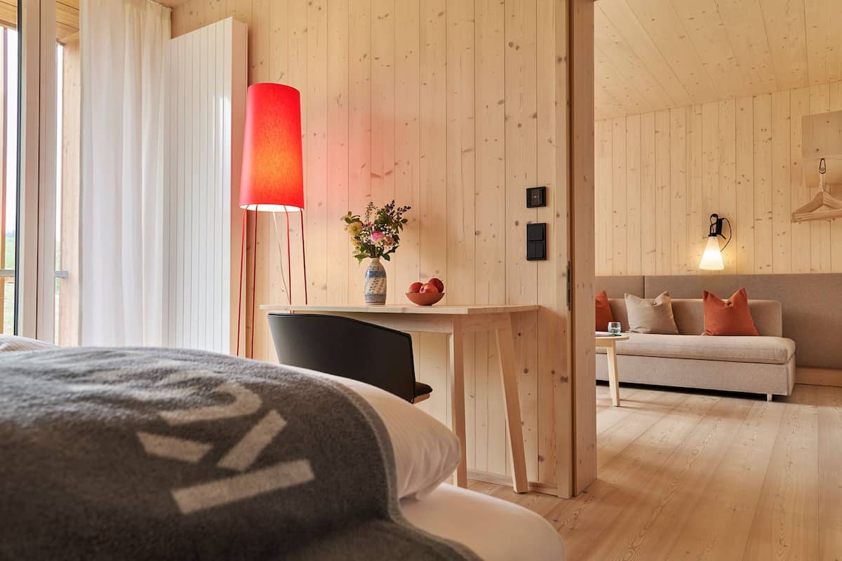 Une chambre d’hôtel moderne avec des murs lambrissés en bois, un lit recouvert d’une couverture grise, un bureau avec une lampe rouge, un vase rempli de fleurs et un canapé agrémenté de coussins orange.
