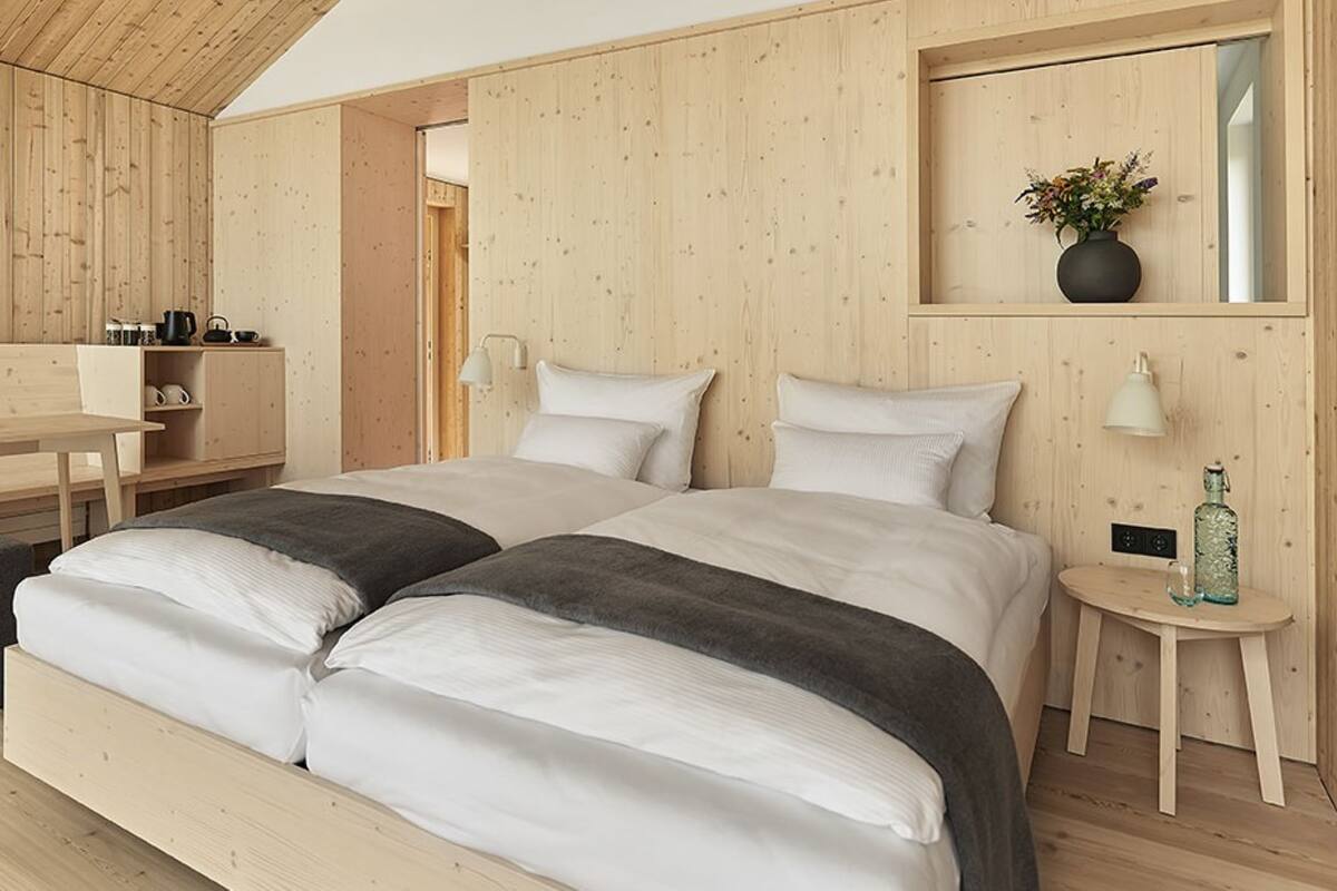 Une chambre moderne avec un décor intérieur en bois, un lit recouvert de draps blancs, une petite table de chevet et un coin bureau agrémenté d’un panier de fruits.