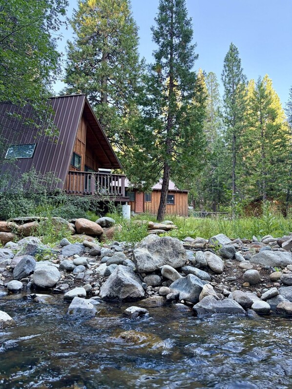 A cabin with a metal roof and wooden deck, surrounded by a rocky stream and tall pine trees.