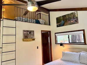 A room with a bunk bed, a ladder, a picture of a forest, a window with a view, and a door. - Forest A-Frame Cabin | Gold Run Creek Frontage | High-Speed WiFi | 10 Wooded Acres (Susanville)