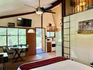 A bedroom with a bed, a desk with chairs, a television, and a kitchen area with an oven and cabinets.