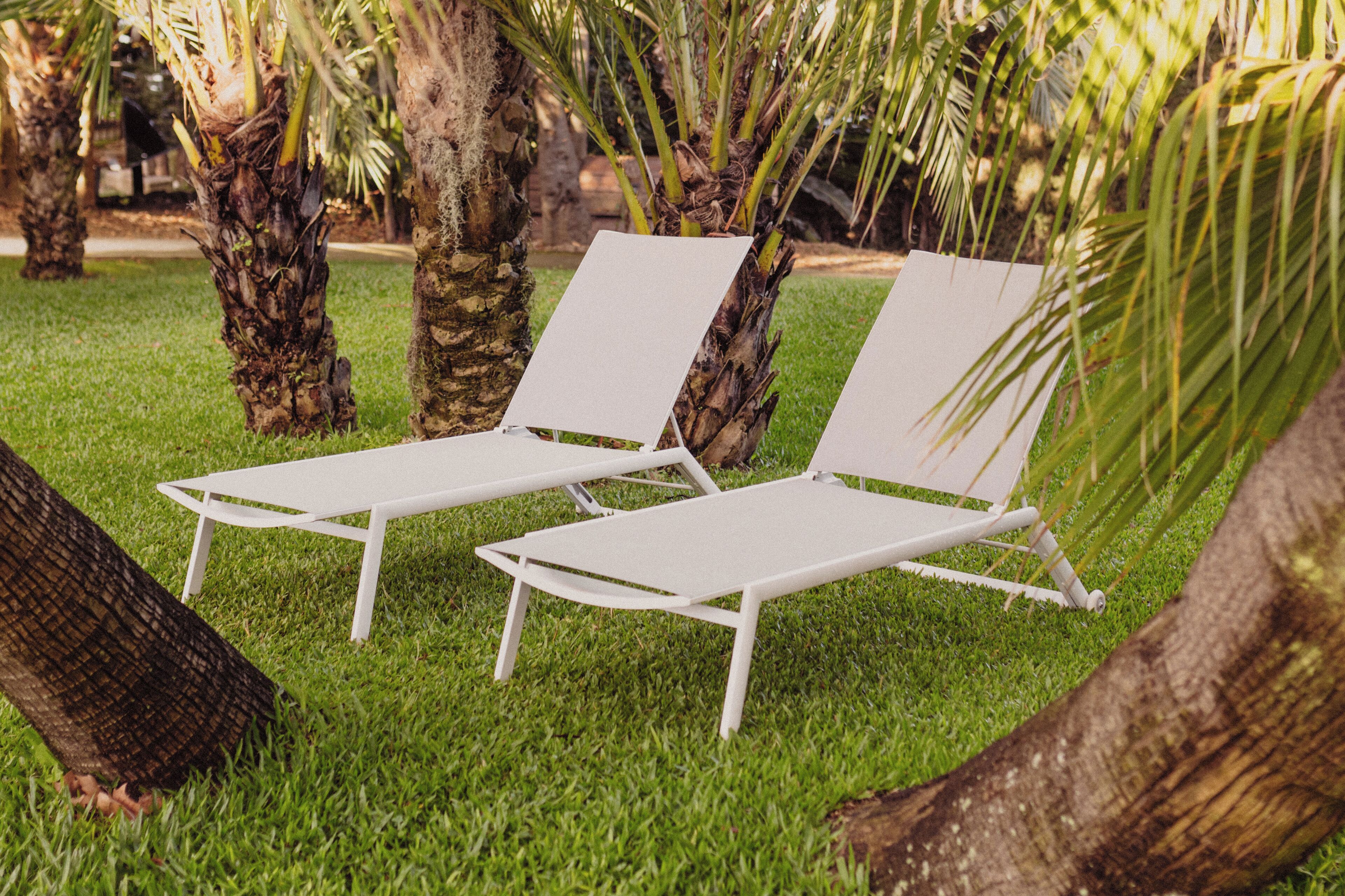 Two white lounge chairs with metal frames, positioned on a well-maintained grassy area with palm trees in the background.