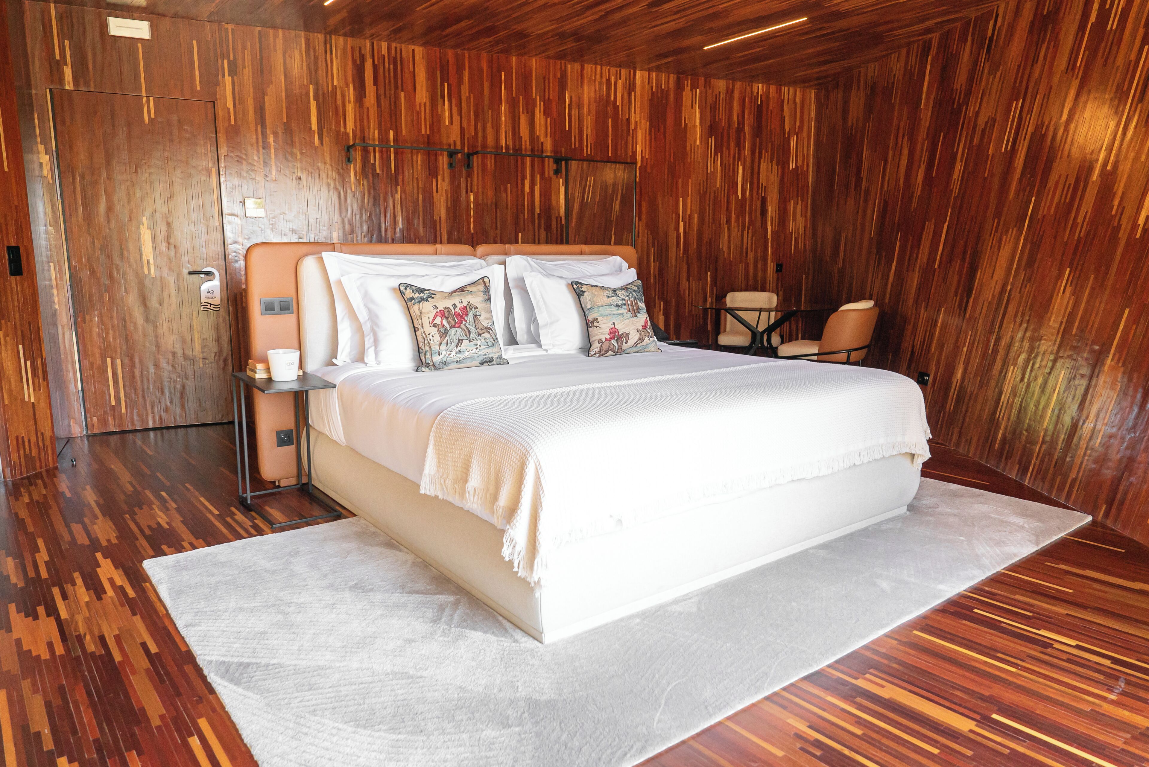 Quarto moderno com uma cama grande, paredes de madeira, um tapete branco e uma pequena mesa com cadeiras.
