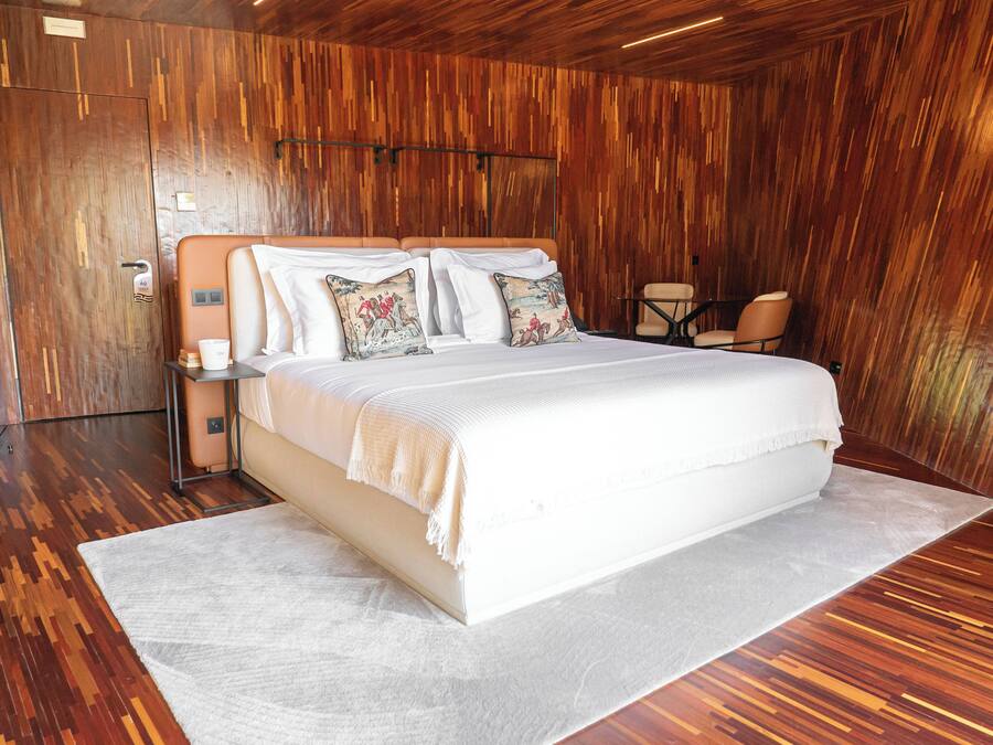 Quarto moderno com uma cama grande, paredes de madeira, um tapete branco e uma pequena mesa com cadeiras.