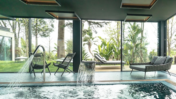 An indoor swimming pool with a waterfall feature, surrounded by lounge chairs and large windows offering views of a tropical garden. - Quinta de Silvalde (Santo Tirso)