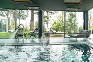 Una piscina interior con una cascada, rodeada de sillas de descanso y grandes ventanales con vistas a un jardín tropical.