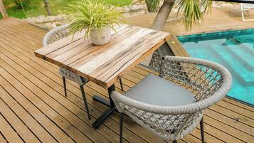 Terraza de madera con mesa y sillas de mimbre, con vistas a una piscina.