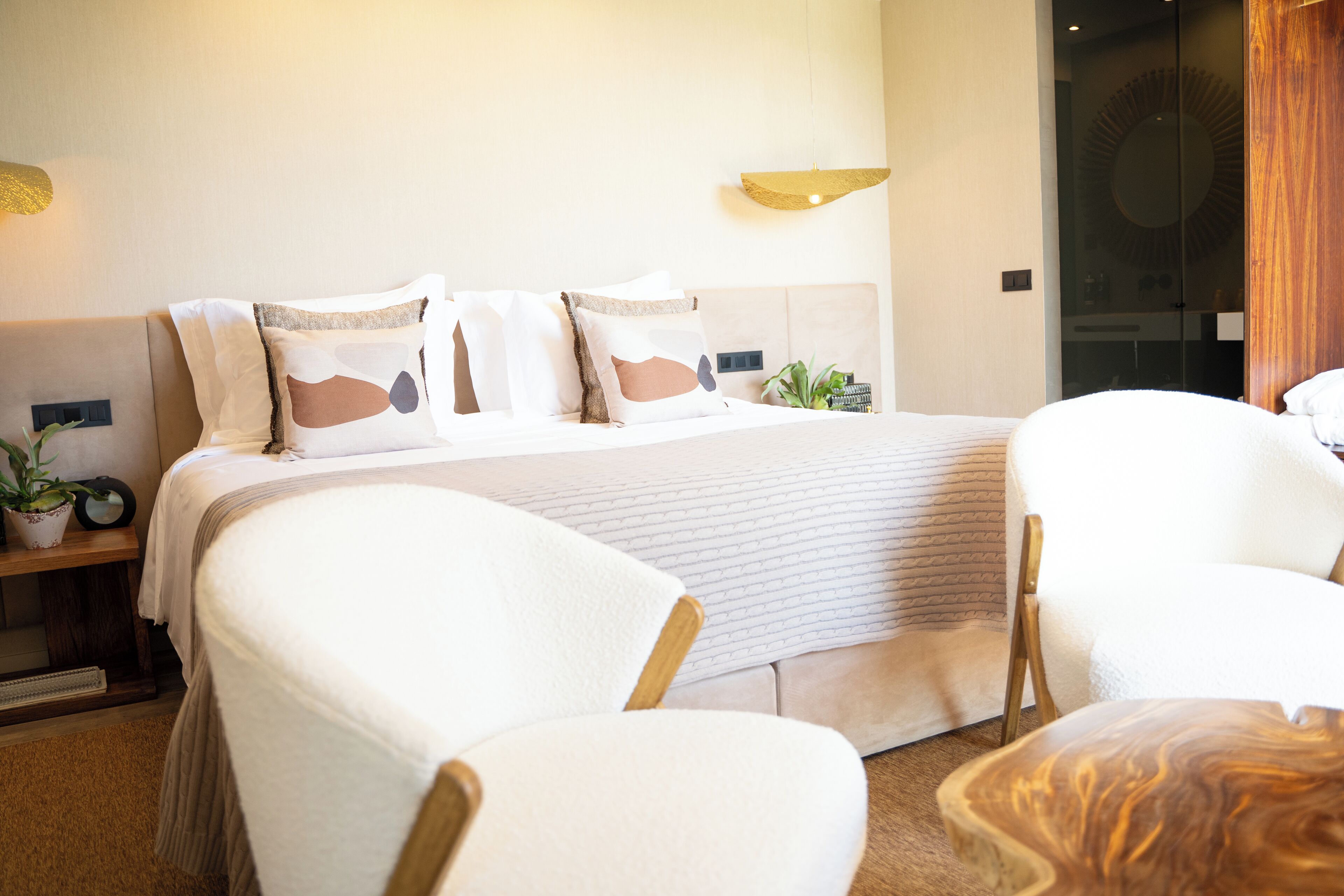 A modern hotel room with a large bed, two white armchairs, a wooden coffee table, and a small side table with a plant.
