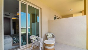 En balkon med to fletstole, et rundt bord og en skydedør, der fører til et soveværelse med en seng og et natbord.