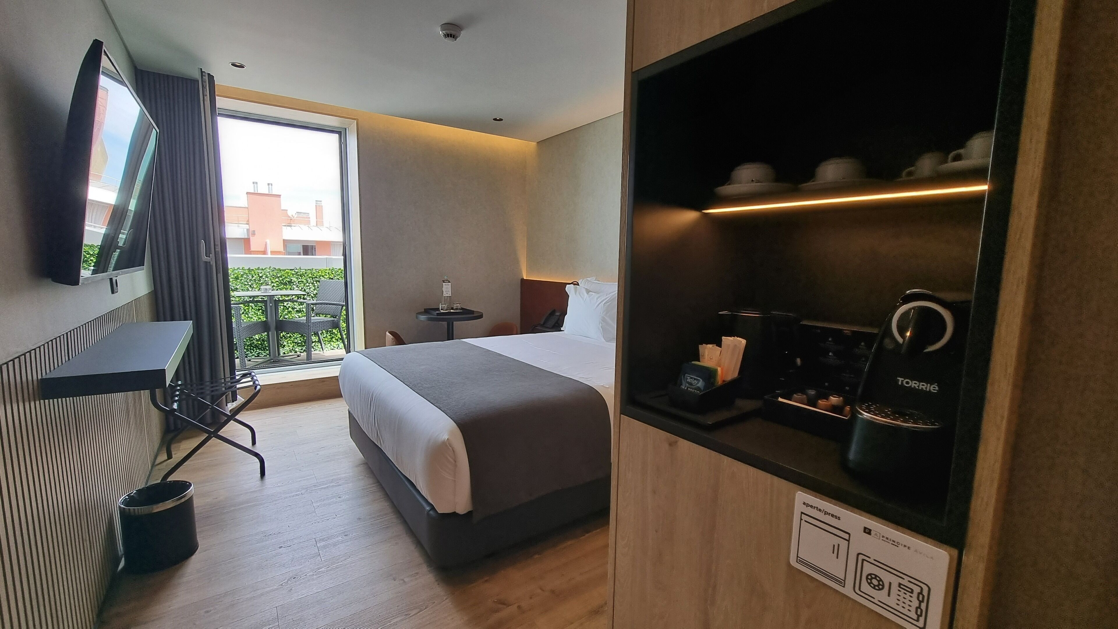 A hotel room with a bed, a desk with a chair, a television, a coffee machine, and a window with a view of buildings.