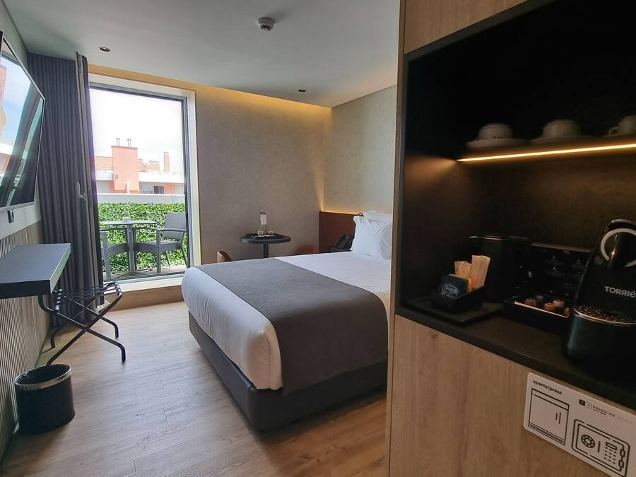 A hotel room with a bed, a desk with a chair, a television, a coffee machine, and a window with a view of buildings.