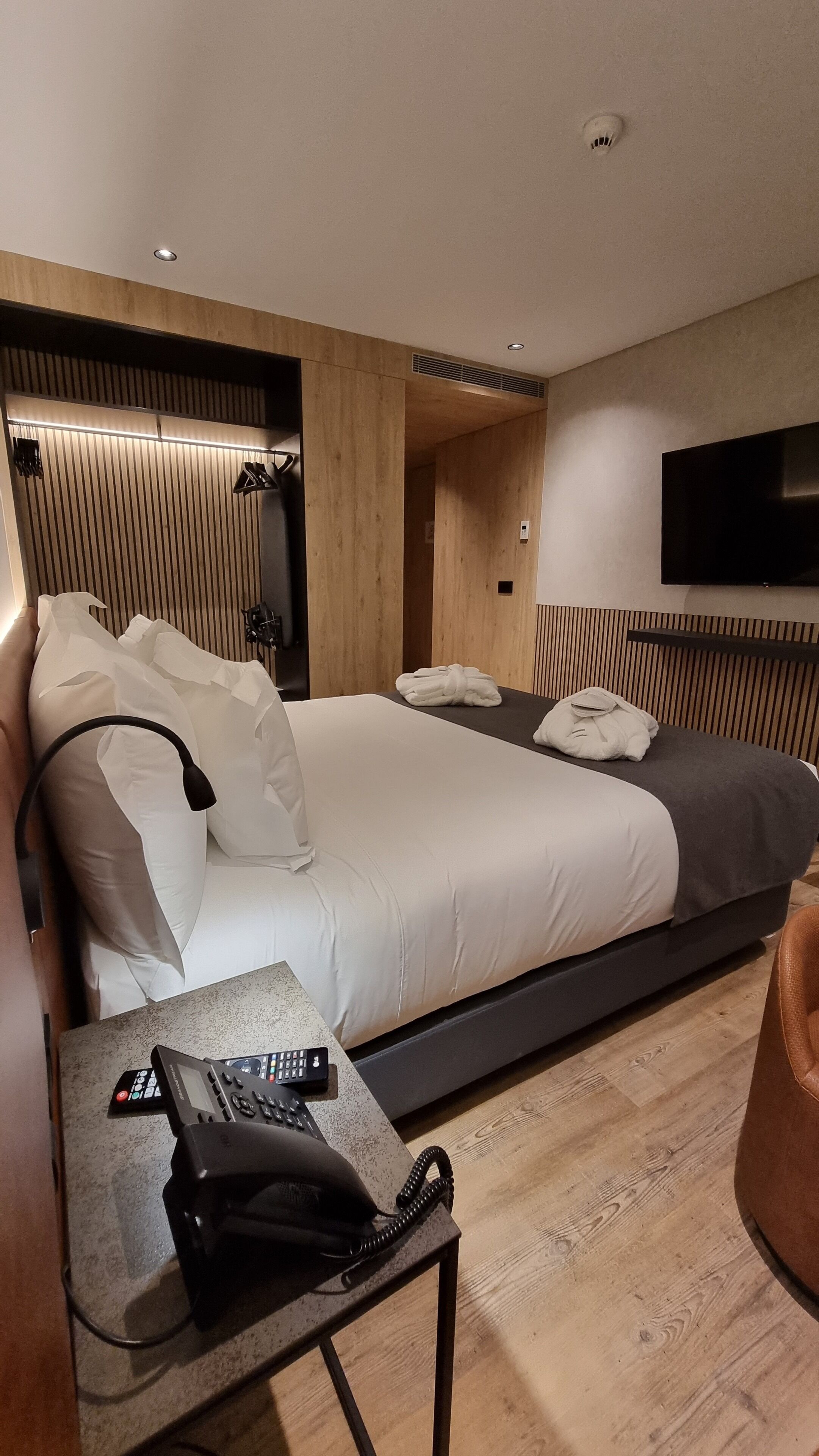 Quarto de hotel moderno com uma cama grande, mesa de cabeceira com telefone e televisão de tela plana na parede.