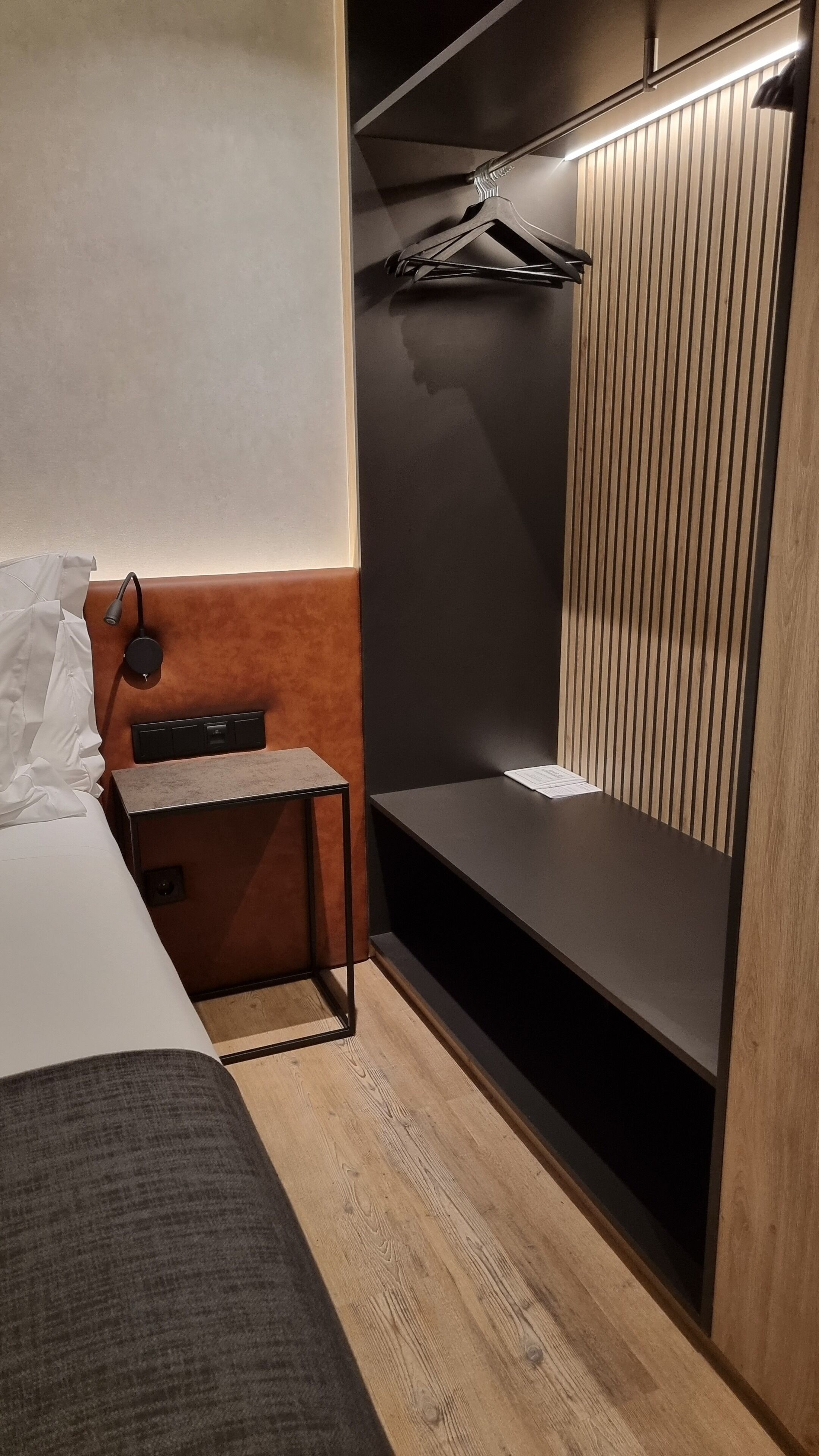 Habitación de hotel pequeña con cama, una mesita, un banco y un armario con perchero.