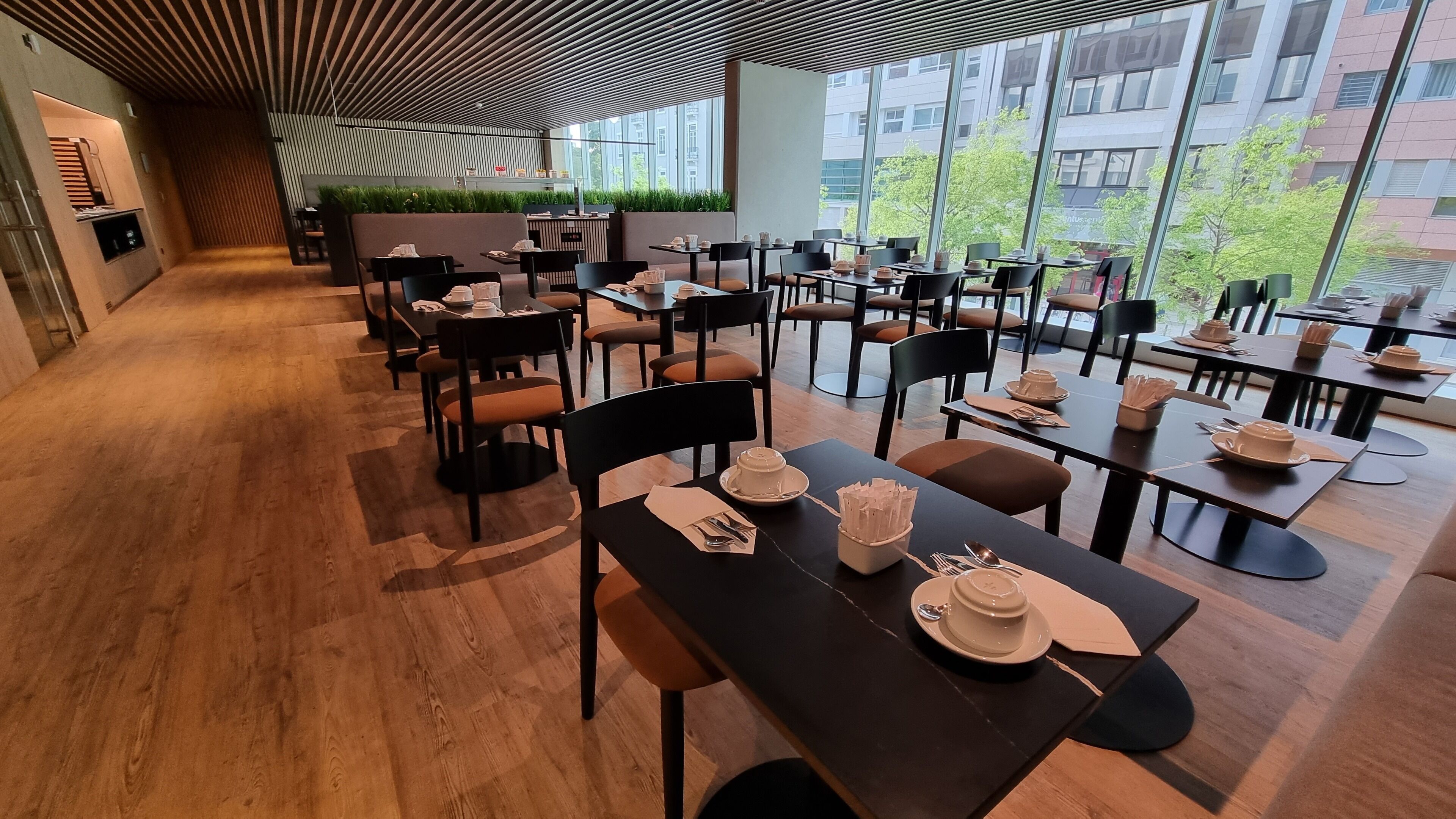 Een modern restaurant met een lange gang naar de eetgedeeltes, grote ramen met uitzicht op de stad, en netjes opgemaakte tafels met stoelen.
