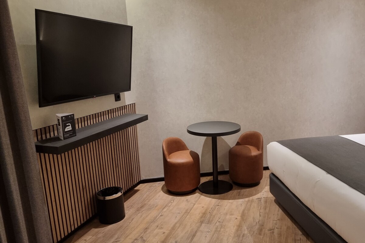 Quarto de hotel com uma cama, televisão fixada na parede, mesa redonda pequena com duas cadeiras, lixeira e piso de madeira.