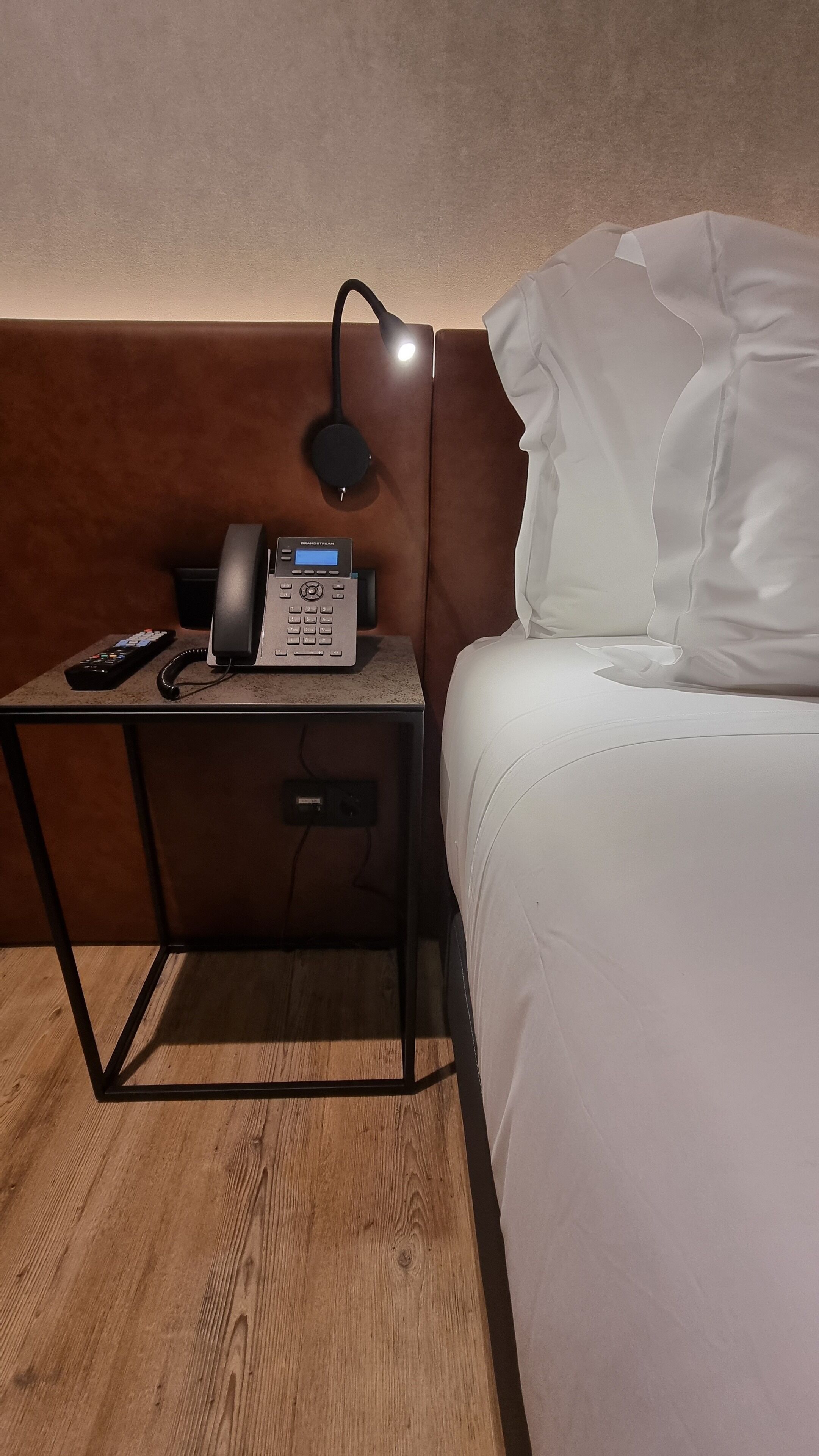 Quarto de hotel com cama, mesa de cabeceira com luminária, telefone e painel de controle.