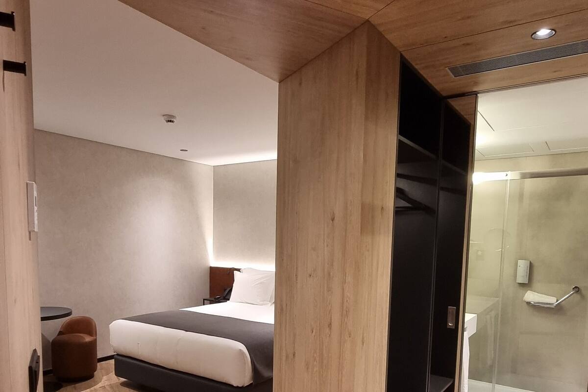 Une chambre d’hôtel moderne avec un plancher en bois, un lit avec une literie blanche, une petite table ronde et une douche en verre.