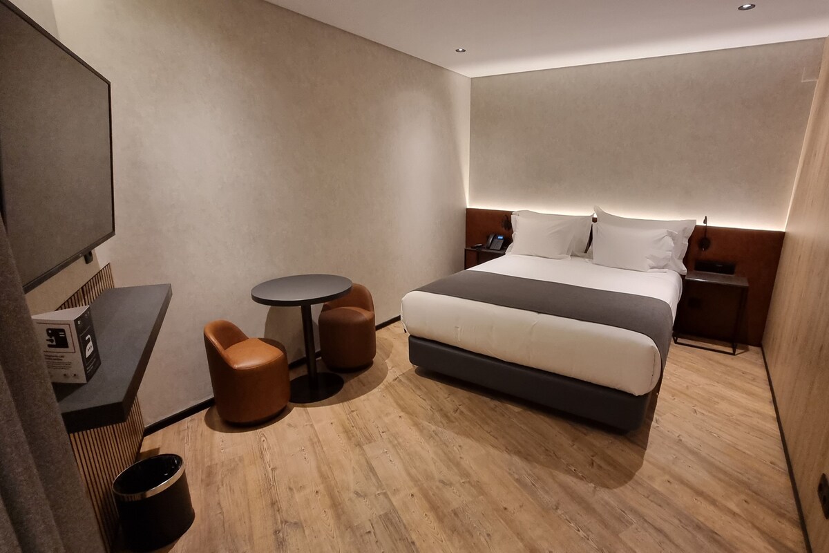 A modern hotel room with a large bed, a small round table, two brown armchairs, a desk with a computer, and a TV.