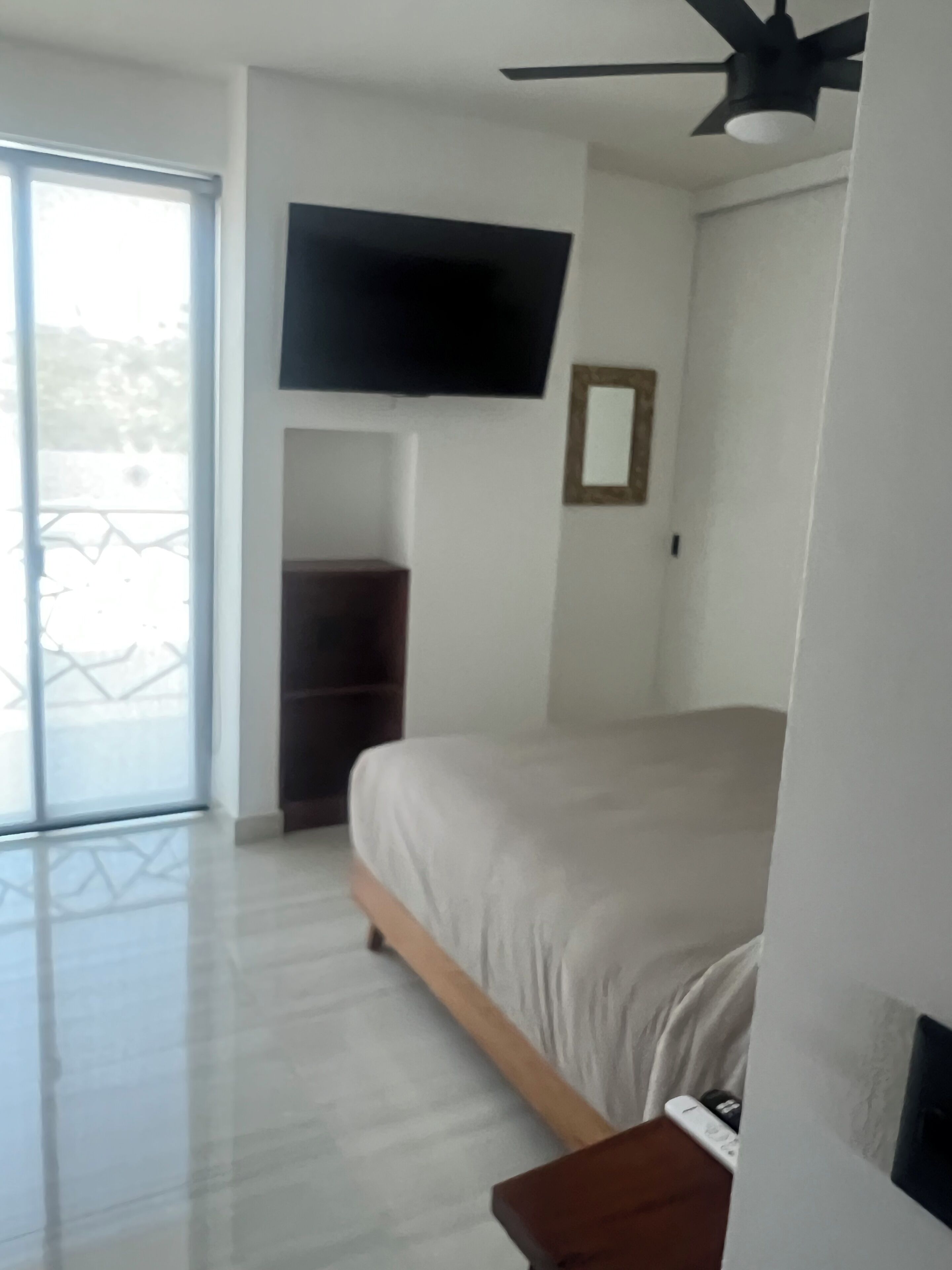 Un dormitorio con cama, televisor de pantalla plana montado en la pared, ventilador de techo y puerta corrediza de vidrio que da a un espacio exterior.