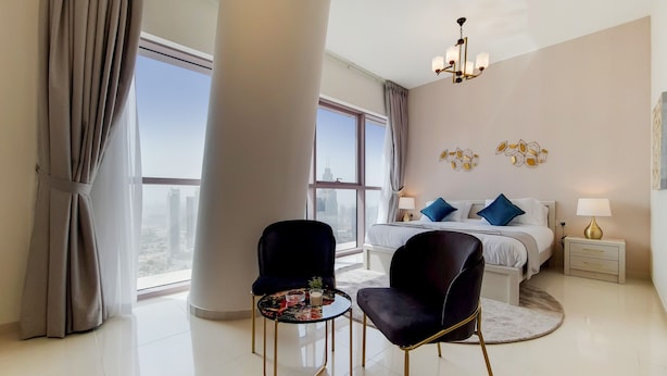 A modern hotel room with a large window, a bed with blue pillows, a round table with chairs, and a nightstand with a lamp.