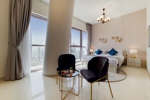 Una habitación de hotel moderna con un ventanal grande, una cama con almohadas azules, una mesa redonda con sillas y una mesita de noche con una lámpara.